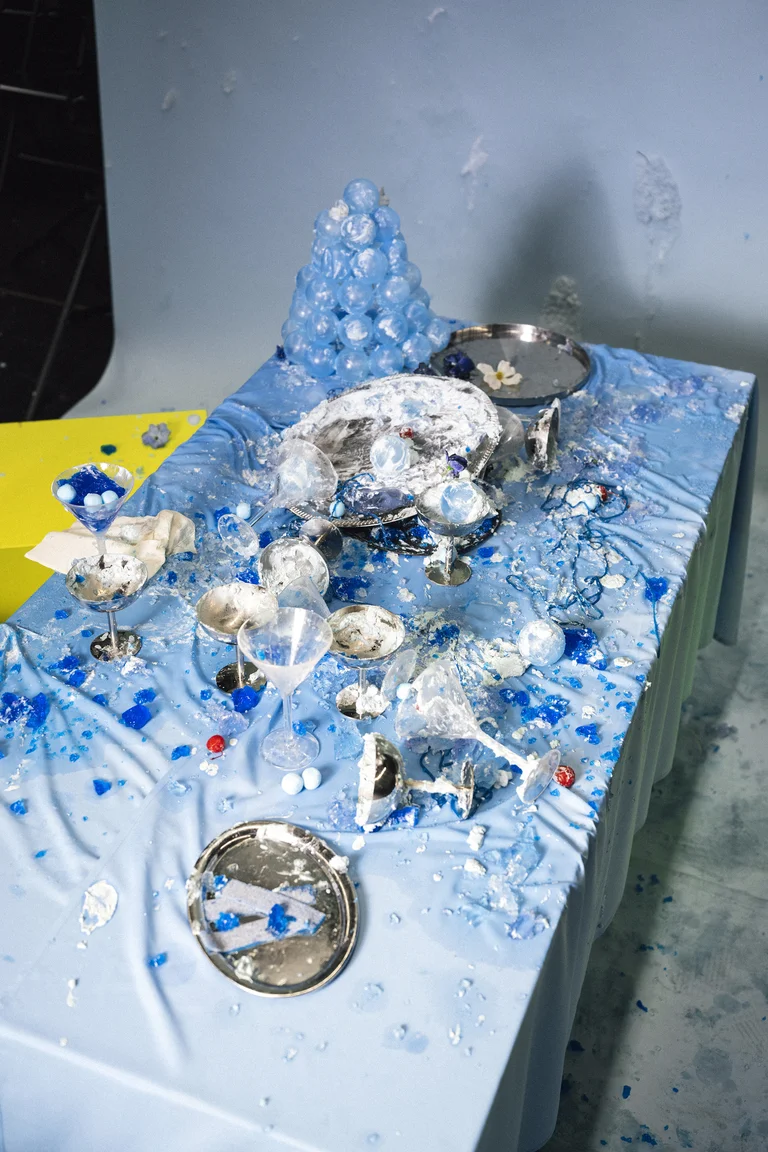 Ein Tisch mit blauer Tischdecke ist mit verschüttetem Essen und zerbrochenem Geschirr bedeckt, umgeben von verstreuten Eiswürfeln.