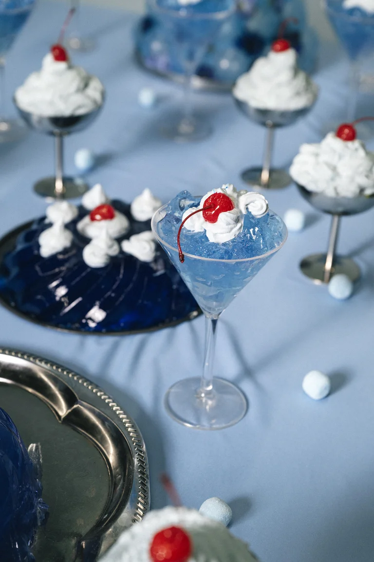 Desserts in eleganten Gläsern auf blauem Hintergrund. Blaue Götterspeise mit Sahnehäubchen und roten Kirschen dekoriert, auf einem Tablett serviert.