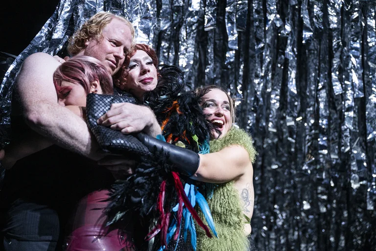 Drei Personen in bunten Kostümen umarmen sich vor einem glitzernden silbernen Hintergrund. Ausdrucksstark und emotionale Performance.