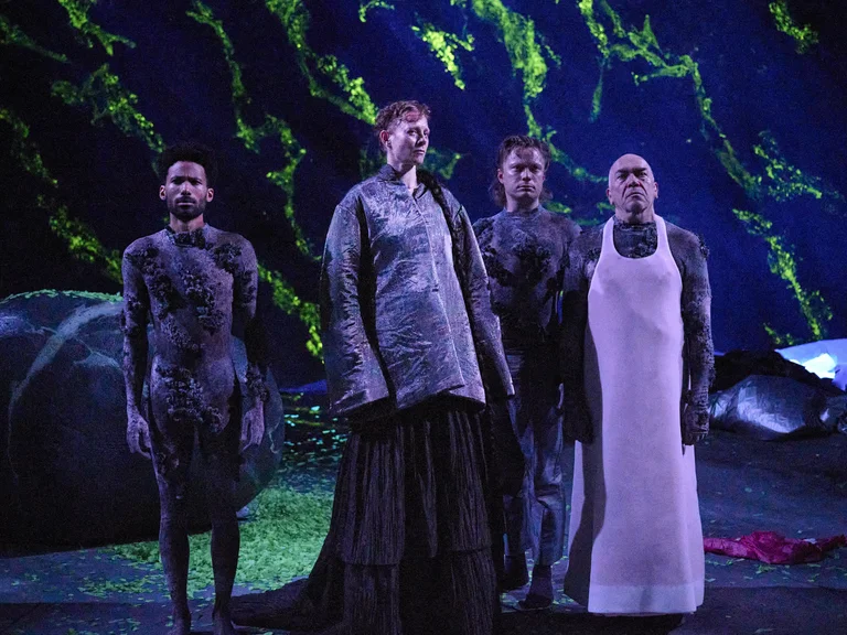 Vier Performer in eindrucksvollen Kostümen stehen auf der Bühne vor einem dramatischen Hintergrund mit grünen und schwarzen Mustern.