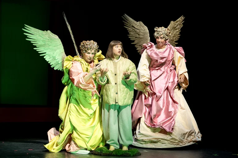Drei Schauspieler auf der Bühne, in bunten, engelhaften Kostümen mit Flügeln. Die Szene wirkt dramatisch und theatralisch inszeniert.