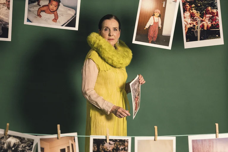 Eine Frau in einem gelben Kleid steht vor einer grünen Wand mit aufgehängten Fotos. Sie hält ein Bild in der Hand.