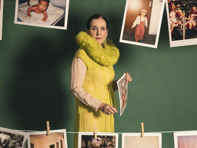 Eine Frau in einem gelben Kleid steht vor einer grünen Wand mit aufgehängten Fotos. Sie hält ein Bild in der Hand.