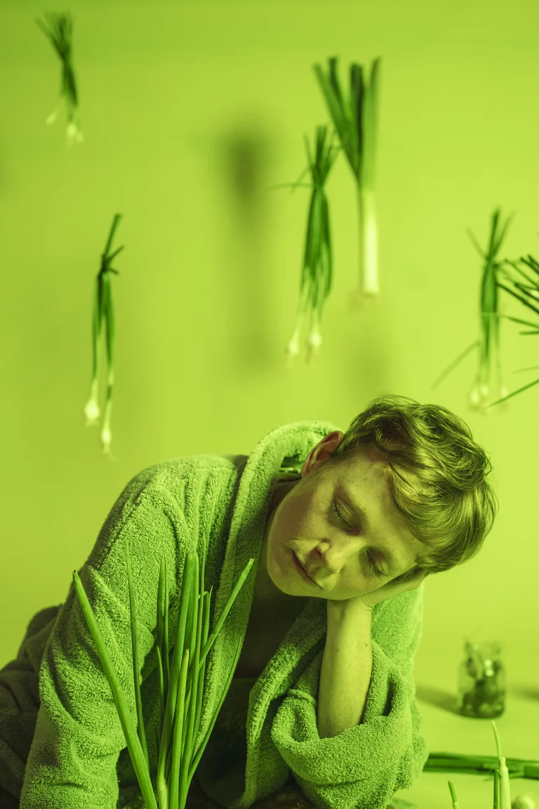 Eine Person in Bademantel ruht auf einem Tisch. Um sie schweben grüne Lauchzwiebeln in einem grün getönten Raum.