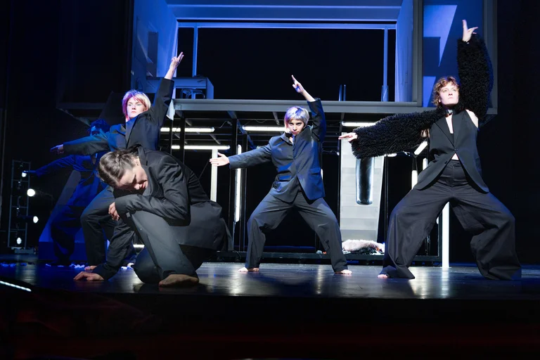 Vier Tänzer in dunklen Anzügen performen energisch auf einer Bühne. Ihre Posen sind dynamisch und ausdrucksstark, wie bei einem modernen Theaterstück.