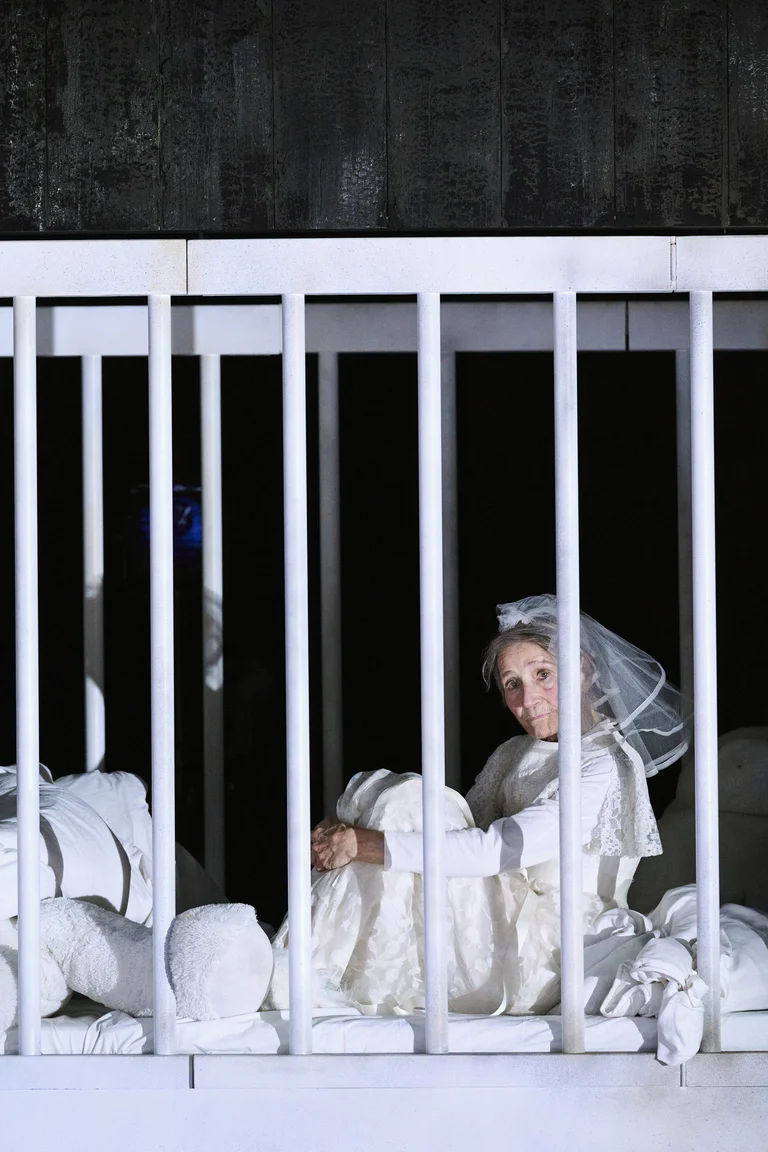 Eine Frau in einem weißen Brautkleid und Schleier sitzt hinter weißen Gitterstäben, umgeben von weißen Stoffen, auf einer dunklen Bühne.