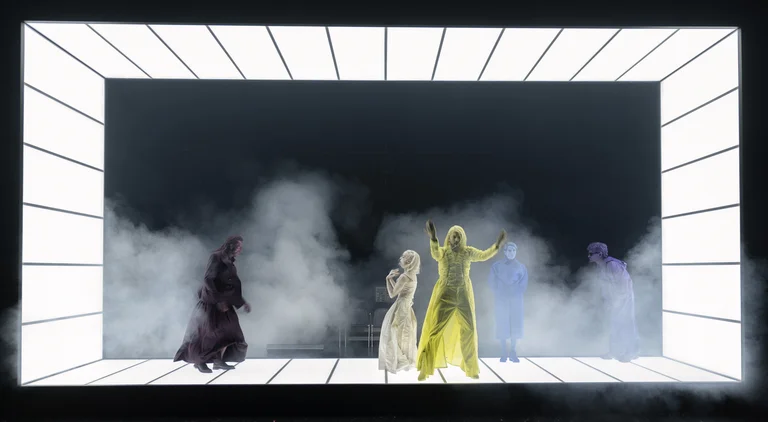 Künstler in leuchtenden Kostümen auf einer futuristischen Bühne, umgeben von Rauch und Nebel, vor einem dunklen Hintergrund.