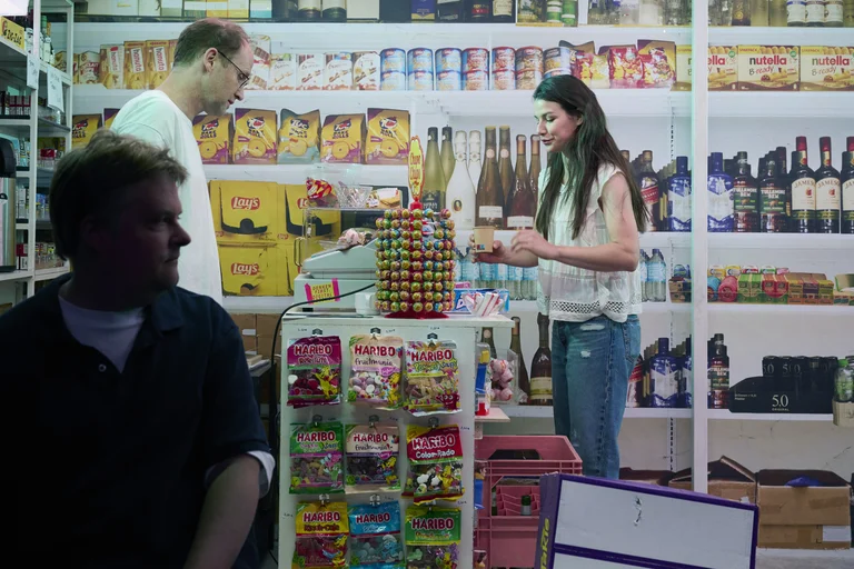 Innenansicht eines kleinen Ladens, eine Frau hinter der Theke bedient einen Kunden. Im Vordergrund sitzt eine Person.