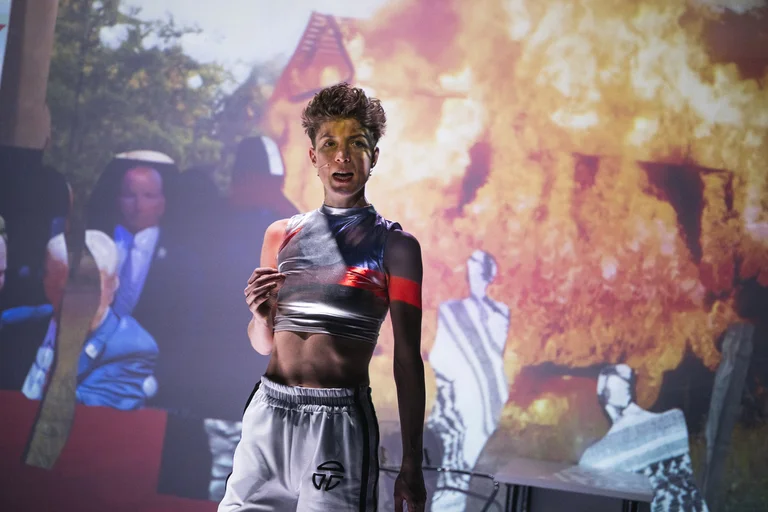 Ein Performer steht vor einer Projektion mit Feuer und Menschen in traditionellen Kleidern. Er trägt sportliche Kleidung.