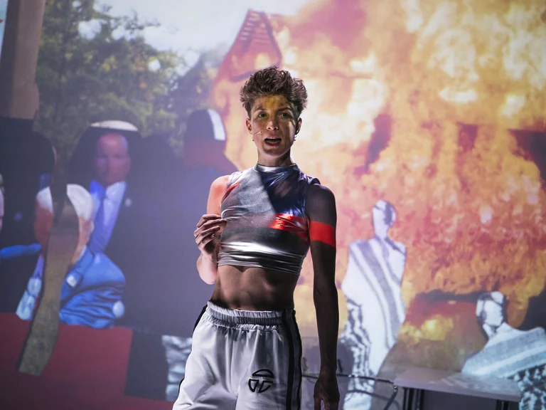 Ein Performer steht vor einer Projektion mit Feuer und Menschen in traditionellen Kleidern. Er trägt sportliche Kleidung.