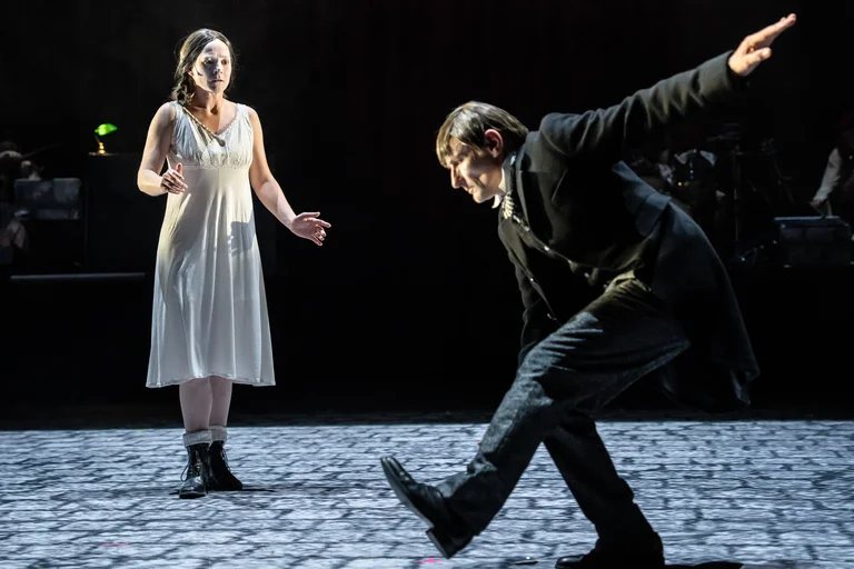 Szene aus einem Theaterstück: Eine Frau in weißem Kleid und Stiefeln steht, während ein Mann in schwarzem Anzug tanzt.