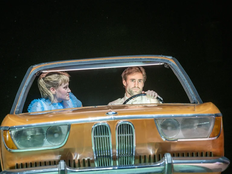 Zwei Personen in einem Auto auf der Bühne, mit dramatischer Beleuchtung und intensiven Ausdrücken, die eine theatralische Szene darstellen.