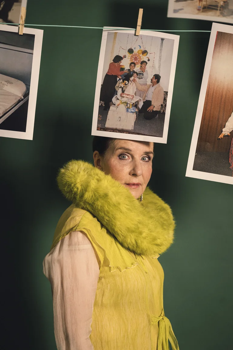 Frau in gelbem Kleid mit flauschigem Kragen; hinter ihr hängen Fotografien an einer Wäscheleine vor grünem Hintergrund.