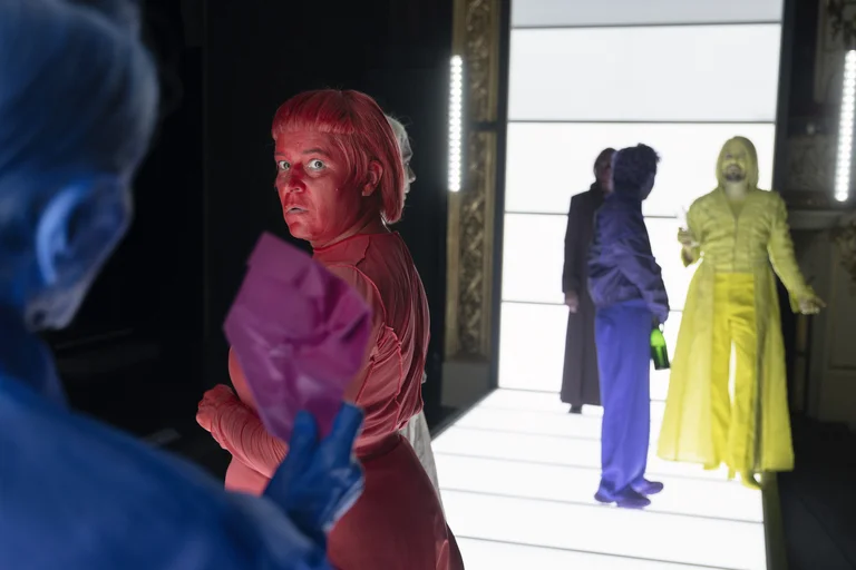 Performancekunst mit Darstellern in bunten, monochromatischen Kostümen und Masken auf beleuchtetem Laufsteg. Farben: blau, rot, lila, gelb.