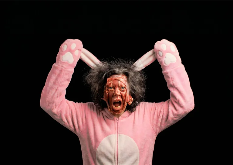 Eine Person in einem rosa Hasenkostüm mit schockierendem Gesichtsausdruck, hält die Hasenohren nach oben. Der Hintergrund ist schwarz.