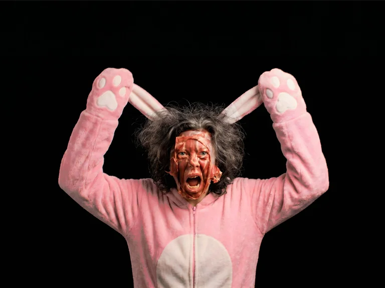 Eine Person in einem rosa Hasenkostüm mit schockierendem Gesichtsausdruck, hält die Hasenohren nach oben. Der Hintergrund ist schwarz.