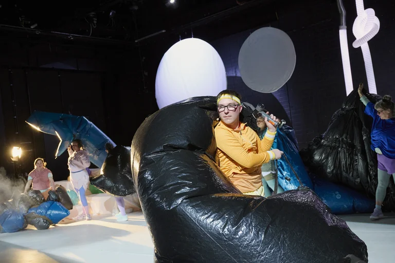 Performers auf der Bühne mit großen aufblasbaren Formen und Lichtspot. Die Szene strahlt Kreativität und lebendige Energie aus.