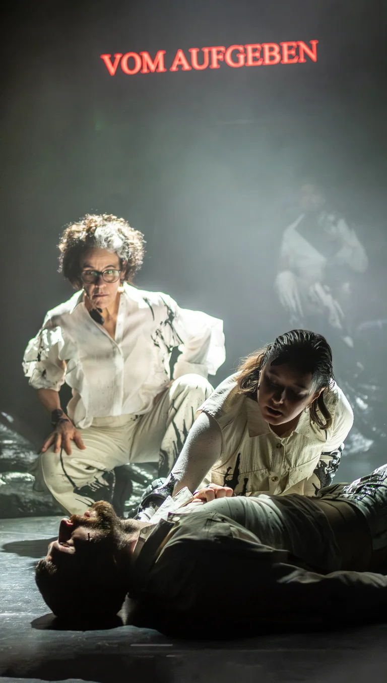 Theaterszene mit zwei Schauspielern, die über einem dritten auf dem Boden gebeugten stehen. Der Text "VOM AUFGEBEN" leuchtet oben.