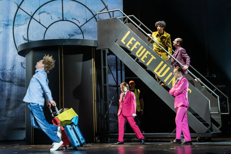 Eine bunte Theateraufführung mit fünf Darstellern in auffälligen Anzügen. Einer zieht einen Koffer die Bühne entlang. Treppe im Hintergrund.