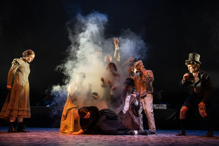 Mehrere Schauspieler in historischen Kostümen führen eine dramatische Szene auf einer Bühne mit Rauch und dunkler Beleuchtung auf.