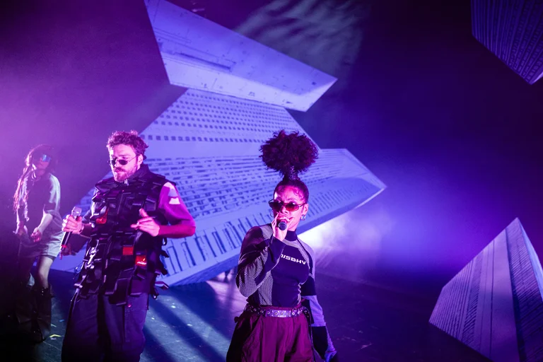 Zwei Musiker auf einer Bühne mit futuristischen Dekorationen und lila Beleuchtung. Die Szenerie wirkt energiegeladen und dynamisch.