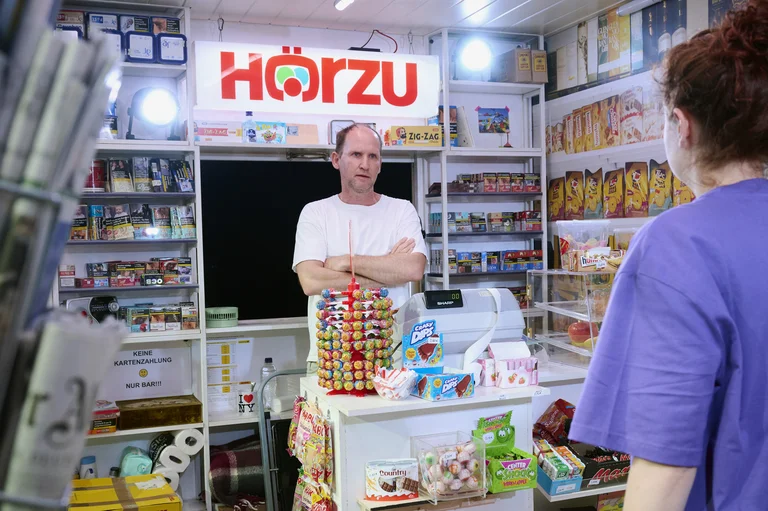 Ein Kioskverkäufer steht hinter der Theke, umgeben von Zeitschriften und Snacks. Ein Schild mit der Aufschrift "Hörzu" leuchtet.