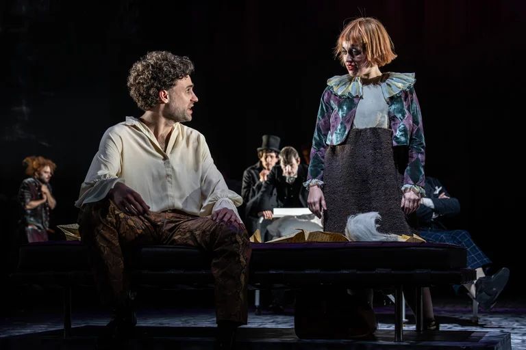 Szene aus einem Theaterstück mit drei Figuren auf der Bühne. Die Atmosphäre wirkt dramatisch und die Kostüme futuristisch.