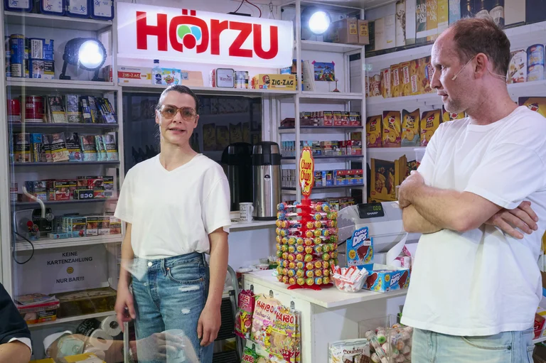 Zwei Personen stehen in einem kleinen Laden, umgeben von Lebensmitteln und Snacks. Ein "Hörzu"-Zeichen leuchtet im Hintergrund.