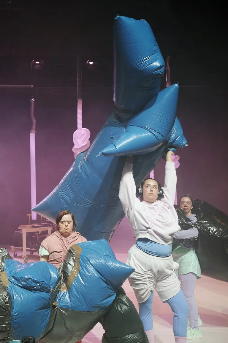 Drei Darstellerinnen in bunten Kostümen heben große, blaue, aufblasbare Objekte in einem theatralischen Bühnenbild mit rosa Licht.