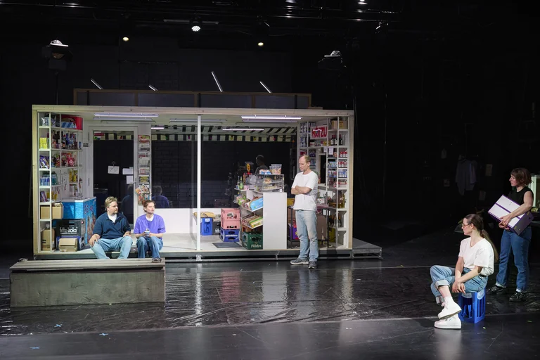 Eine Theaterbühne zeigt eine Kulisse eines kleinen Geschäfts. Mehrere Personen sind unterschiedlich positioniert, zwei sitzen und andere stehen.