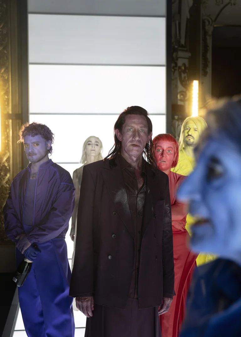Menschen stehen in farbigen Anzügen und mit bemalten Gesichtern in einer Reihe. Der Hintergrund ist in hellem Licht beleuchtet.