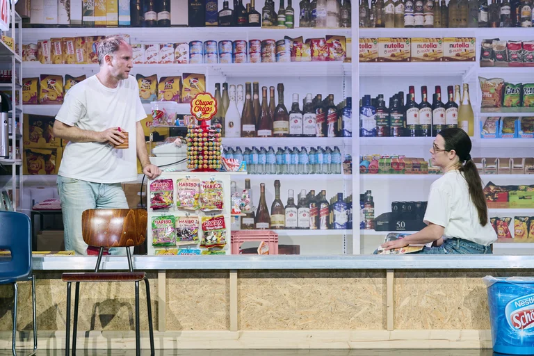 Zwei Personen spielen in einem Bühnenbild, das wie ein Laden aussieht. Regale voller Produkte sind im Hintergrund sichtbar.