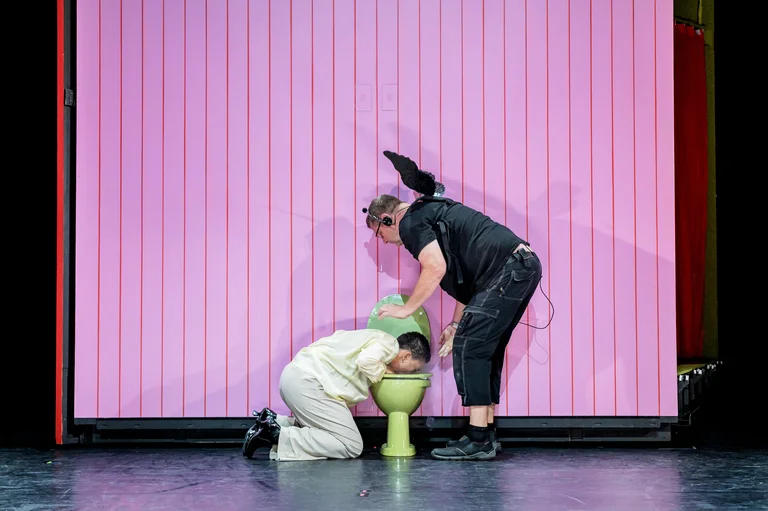 Zwei Performer auf einer Bühne; einer kniet nieder, während der andere eine grüne Requisite hält, vor einem rosa Hintergrund.