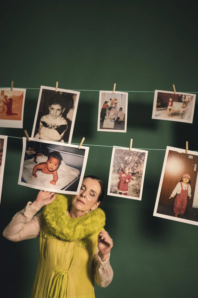 Eine Frau in gelbem Kleid betrachtet Fotos von Kindern an einer Wäscheleine. Die Fotos zeigen verschiedene Lebensabschnitte.