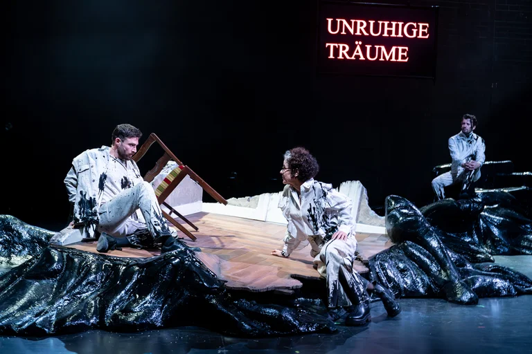 Theaterszene mit drei Personen in weißen Outfits auf einer Bühne mit schwarzem Hintergrund. Der Text "UNRUHIGE TRÄUME" leuchtet rot.