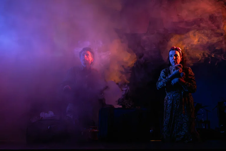 Zwei Performer stehen auf einer Bühne mit dramatischer Beleuchtung, umgeben von farbigem Nebel in Violett- und Orangetönen.