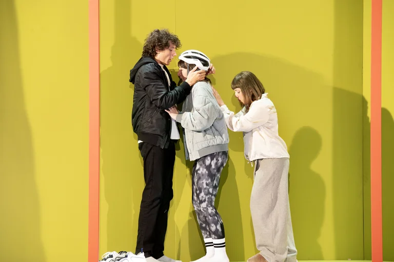 Drei Personen stehen nah beieinander vor einer gelben Wand. Sie scheinen in einer nachdenklichen oder emotionalen Szene zu interagieren.