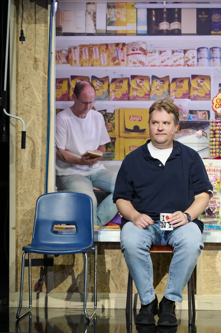 Ein Mann in einem blauen Hemd sitzt auf einem Stuhl mit einer Tasse in der Hand. Hintergrund: ein Plakat eines Supermarkts.