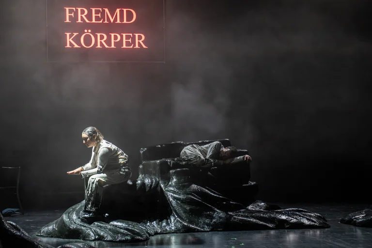Eine Person sitzt auf einer Bühne mit düsterem Hintergrund. Der Schriftzug "Fremd Körper" leuchtet im Hintergrund.