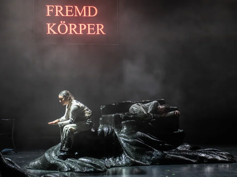 Eine Person sitzt auf einer Bühne mit düsterem Hintergrund. Der Schriftzug "Fremd Körper" leuchtet im Hintergrund.