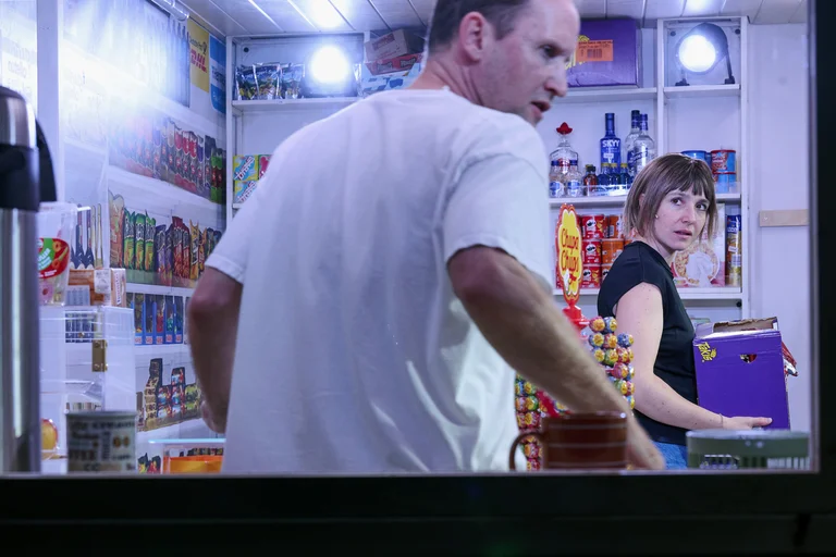 Zwei Personen in einem kleinen Laden, umgeben von Regalen mit Süßigkeiten und Snacks. Eine Person hält ein lila Buch.