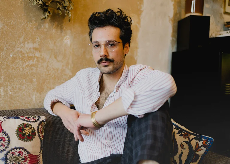 Ein Mann mit Brille und Schnurrbart sitzt lässig auf einem Sofa vor einer rustikalen Wand.