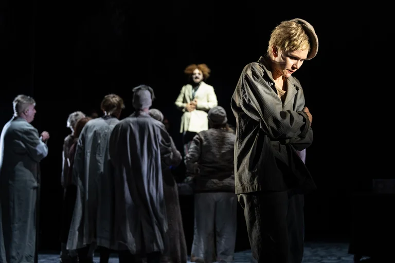 Eine Theaterszene mit mehreren Schauspielern in dunkler Kleidung. Im Hintergrund steht eine Figur mit clownartigem Haar.