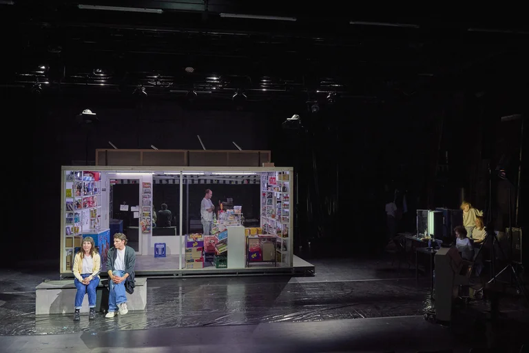 Eine Theaterbühne zeigt eine beleuchtete Bude mit Schauspielern. Die Szene wirkt modern und alltäglich, mit dunkler Atmosphäre.