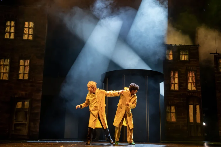Zwei Schauspieler in Trenchcoats performen auf einer düsteren Bühne mit Stadtbild und dramatischer Beleuchtung. Geheimnisvolle Atmosphäre.
