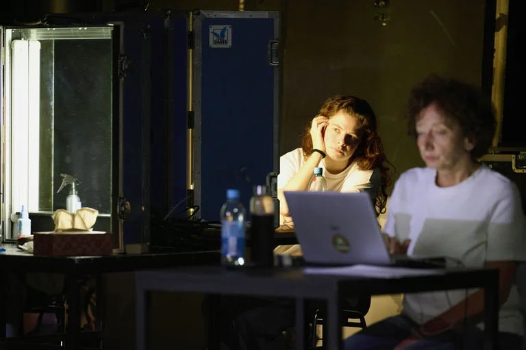 Zwei Personen sitzen an einem Tisch in einem dunklen Raum, beide vertieft in ihre Laptops. Eine Flasche Wasser steht da.