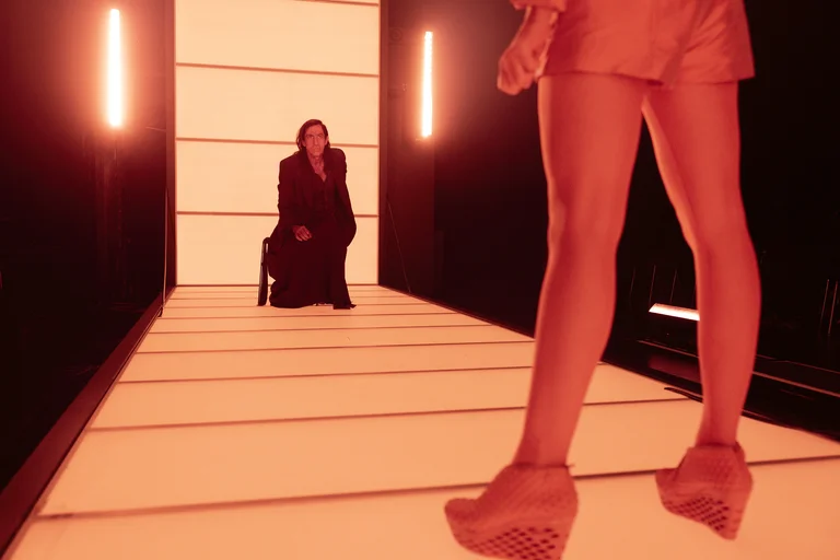 Zwei Personen in einem futuristisch beleuchteten Gang. Eine sitzt in Schwarz, die andere steht in modischer Kleidung und hohen Schuhen.