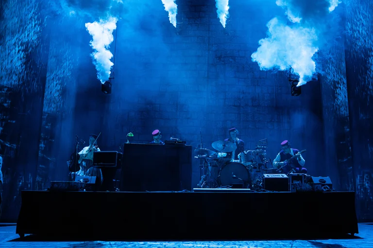 Eine Bühne in blauer Beleuchtung mit einer Band, die ein Live-Konzert gibt. Rauchwolken steigen dramatisch in den Hintergrund auf.