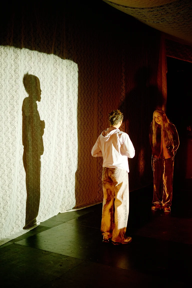 Zwei Performer stehen vor einer Wand, beleuchtet, so dass ihre Schatten sichtbar sind. Die Szene erzeugt eine theatralische Atmosphäre.