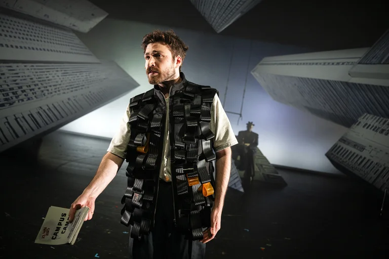 Ein Schauspieler steht auf einer Bühne mit futuristischen Hintergrundprojektionen. Er trägt eine Weste mit vielen Taschen.